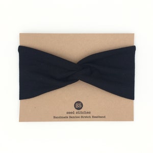 May include: Black headband made from bamboo fabric. The headband is twisted and has a brown paper tag with the text "seed stitches" and "Handmade Bamboo Stretch Headband".