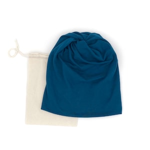 Puede incluir: Un gorro de punto azul turquesa holgado con un nudo en la parte superior, empaquetado en una bolsa de lona natural con cordón.