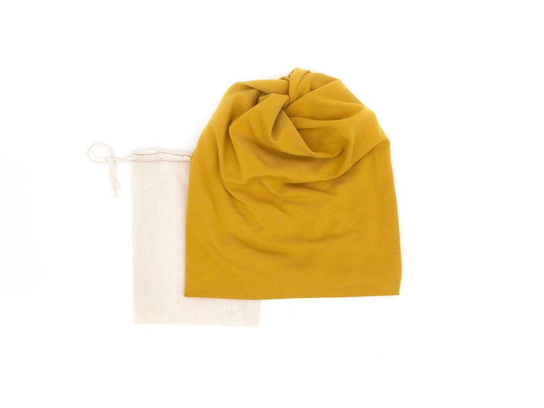 Pu&ograve; includere: Un berretto giallo senape e una borsa in lino naturale con coulisse. Il berretto &egrave; realizzato in un tessuto morbido ed elastico e ha una cima annodata.
