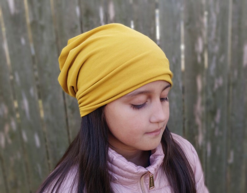 Pu&ograve; includere: Un berretto soffice in tessuto giallo senape, con un design slouchy. Il berretto &egrave; indossato da una persona con capelli lunghi e castani.