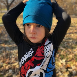 Pu&ograve; includere: Un bambino indossa un berretto blu turchese e una maglietta nera a maniche lunghe con una stampa grafica del logo e dei personaggi degli Avengers. La maglietta ha la scritta "Avengers" stampata.