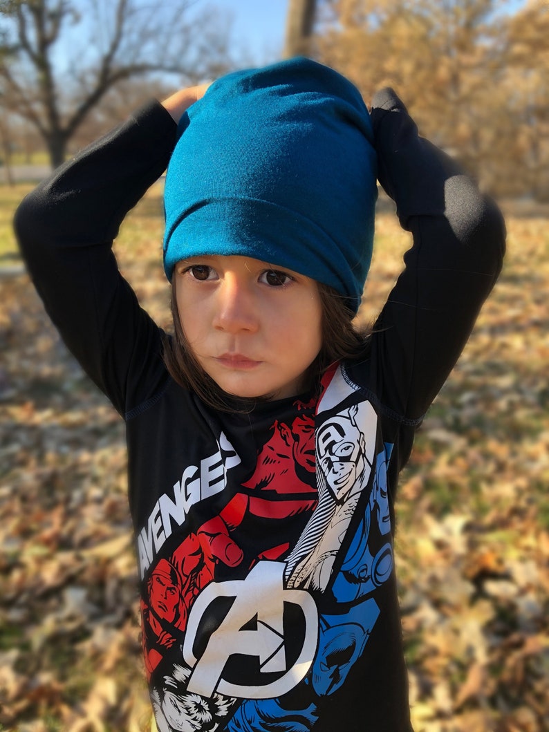 Pu&ograve; includere: Un bambino indossa un berretto blu turchese e una maglietta nera a maniche lunghe con una stampa grafica del logo e dei personaggi degli Avengers. La maglietta ha la scritta "Avengers" stampata.