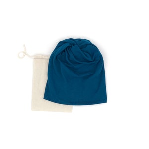 Pu&ograve; includere: Un berretto in tessuto blu turchese con un nodo sulla parte superiore, piegato e posizionato sopra una borsa con coulisse beige.