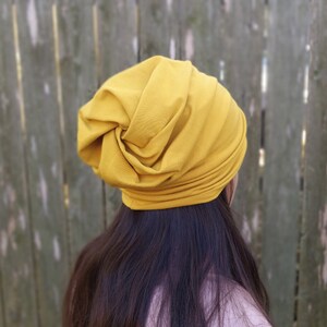 Pu&ograve; includere: Un cappello in stile turbante in tessuto morbido di colore senape. Il cappello &egrave; indossato sulla testa ed &egrave; stilizzato con un nodo sulla parte superiore.
