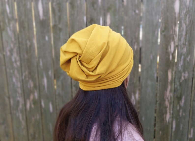 Pu&ograve; includere: Un cappello in stile turbante in tessuto morbido di colore senape. Il cappello &egrave; indossato sulla testa ed &egrave; stilizzato con un nodo sulla parte superiore.