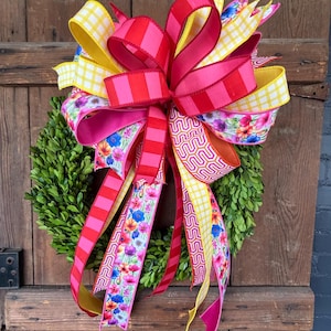 May include: A vibrant wreath with a lush green base, adorned with a large bow. The bow features ribbons in various patterns: red stripes, yellow gingham, and floral prints. The wreath is displayed against a rustic wooden door.