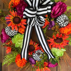 May include: A vibrant autumn wreath featuring sunflowers, orange mums, and purple flowers. The wreath is adorned with small, black and white checkered pumpkins and a large black and white striped bow. Green foliage adds texture.