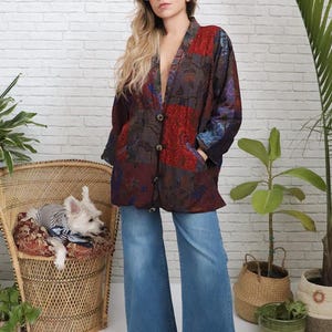 May include: A patterned patchwork jacket in red, brown, and blue with a floral design. The jacket has a button closure and pockets. The model is wearing blue denim jeans and brown boots. A small dog is in a wicker chair.