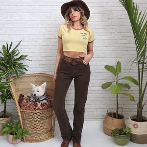 Puede incluir: Una mujer con un top corto amarillo con ribetes marrones y texto, pantalones marrones, un cinturón marrón y un sombrero marrón. Un perro blanco está sentado en una silla de mimbre. El fondo es una pared de ladrillos blancos con plantas.