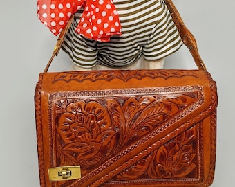 May include: A white dog wearing a striped shirt and a red polka dot scarf, holding a brown leather handbag with floral tooling. The bag has a gold-colored clasp and a long strap.