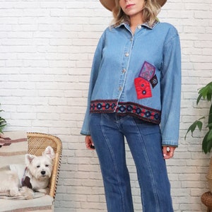 May include: A light blue denim jacket with colorful embroidered trim and patches. The jacket has a button closure and long sleeves. The model is wearing the jacket with a brown hat and blue jeans.