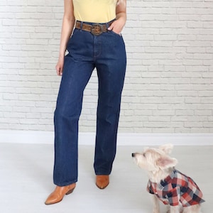 May include: A woman wearing a yellow t-shirt with a brown belt and dark blue jeans. She is wearing brown cowboy boots. A small white dog wearing a red and black checked shirt is sitting in front of her.