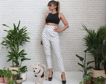 May include: A woman wearing a black tank top and white polka dot trousers stands in front of a white brick wall. She is wearing black sunglasses and black heels. A small white dog is sitting in front of her.