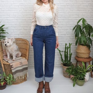 K&ouml;nnte beinhalten: Eine Frau tr&auml;gt einen wei&szlig;en Strickpullover und eine blaue, hochgeschlossene Jeans mit umgeschlagenen S&auml;umen. Sie steht vor einer wei&szlig;en Backsteinwand mit Pflanzen und einem Korbstuhl.
