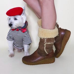 May include: A white dog wearing a red beret, a striped shirt, and a red bow tie. The dog is standing next to a person wearing brown boots with a tan lining and a brown and tan skirt.