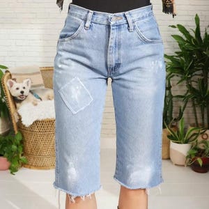 Pode incluir: Calças capri de ganga azul claro com bainha crua e um remendo quadrado na perna esquerda. As calças têm salpicos de tinta branca e são usadas com um top preto e um lenço estampado.