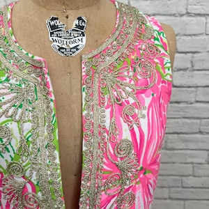 May include: A sleeveless tunic dress featuring a vibrant pink, green, and white floral pattern. The garment has a deep split neckline adorned with intricate metallic gold embroidery that outlines the collar and the front placket edges.