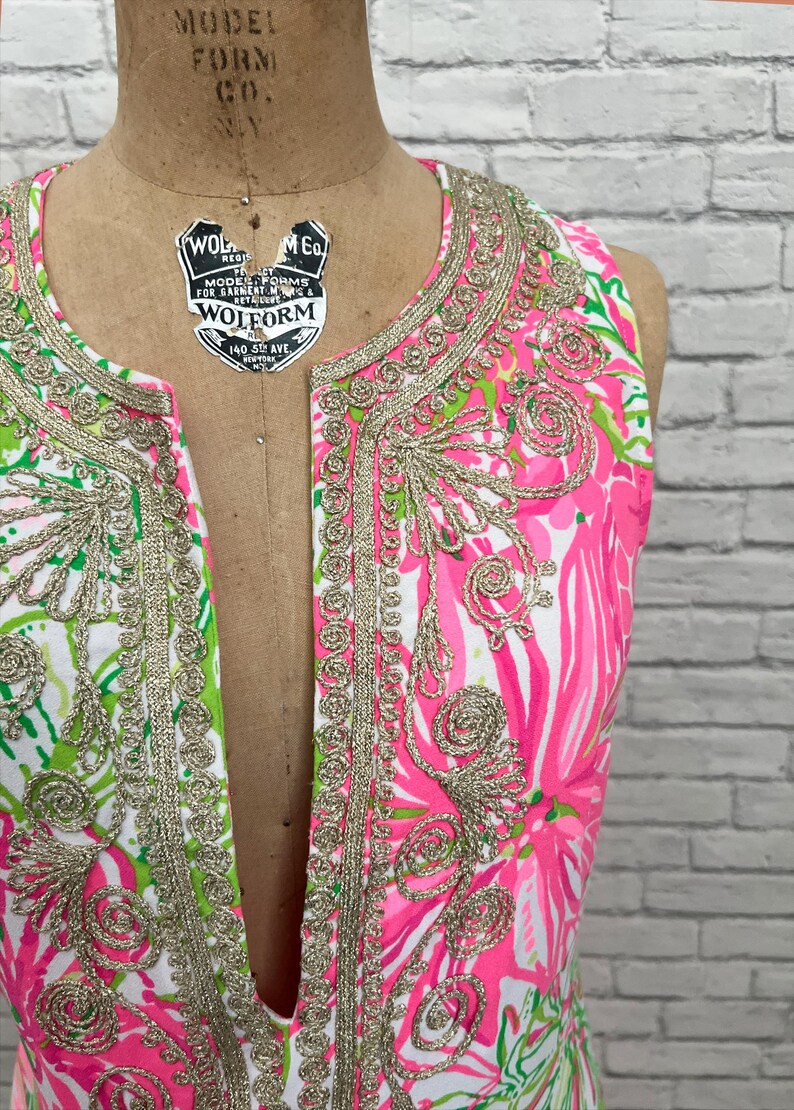 May include: A sleeveless tunic dress featuring a vibrant pink, green, and white floral pattern. The garment has a deep split neckline adorned with intricate metallic gold embroidery that outlines the collar and the front placket edges.