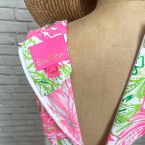 May include: A sleeveless dress featuring a vibrant pink, lime green, and white floral pattern. The garment includes a white zipper closure along the back and a bright pink interior label with the text Lilly Pulitzer and the size 4 printed in gold.