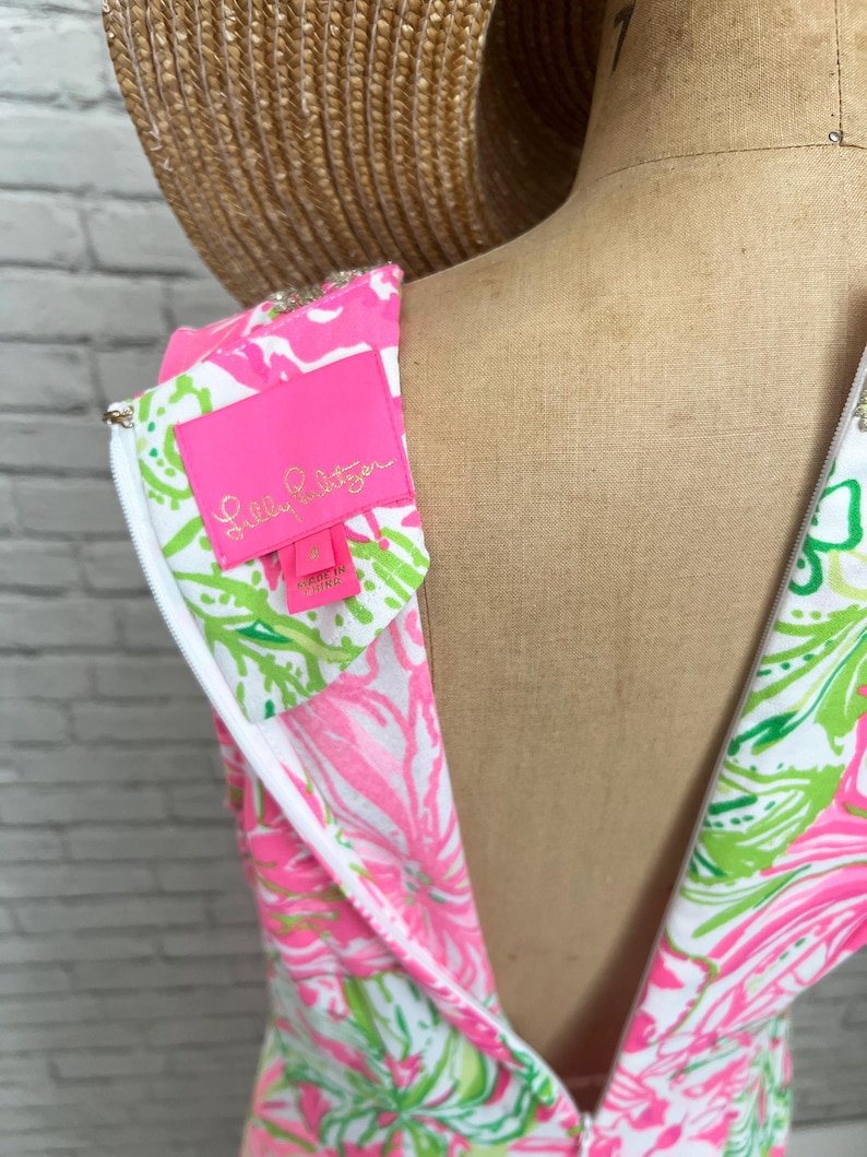 May include: A sleeveless dress featuring a vibrant pink, lime green, and white floral pattern. The garment includes a white zipper closure along the back and a bright pink interior label with the text Lilly Pulitzer and the size 4 printed in gold.
