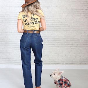 May include: A woman wearing a yellow t-shirt with the text "Do it in the canyon" on the back, blue jeans, a brown cowboy hat, and brown boots. A small white dog wearing a red and black plaid shirt is sitting next to her.