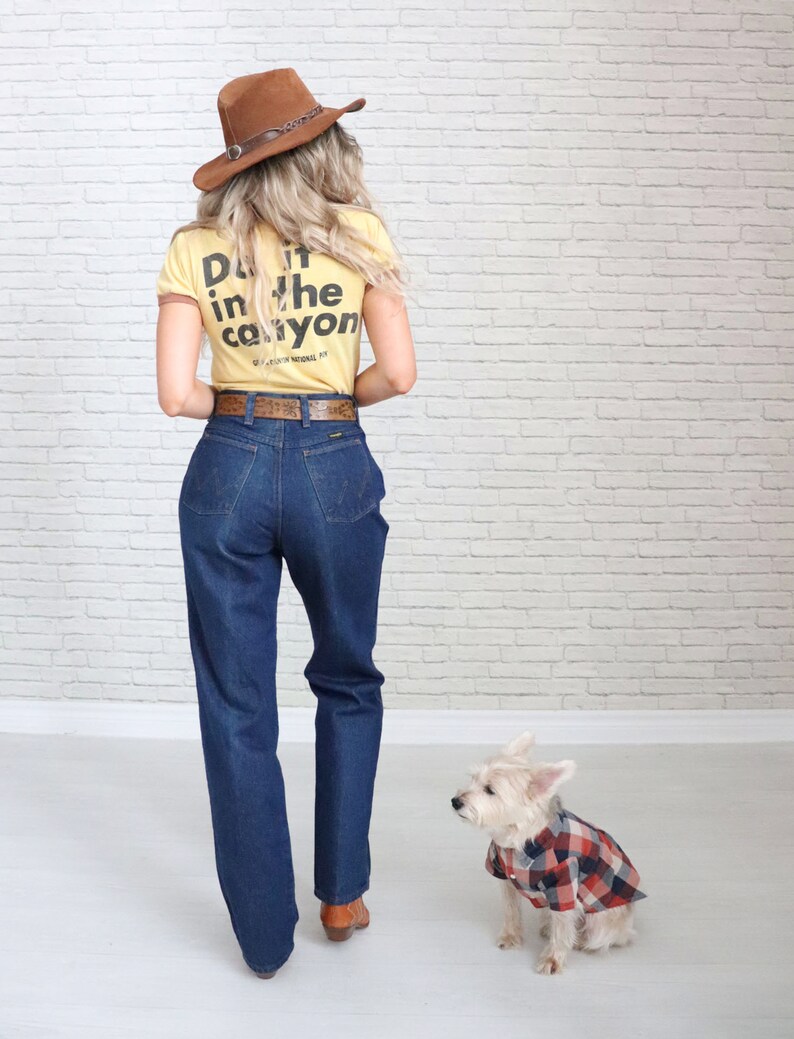 May include: A woman wearing a yellow t-shirt with the text "Do it in the canyon" on the back, blue jeans, a brown cowboy hat, and brown boots. A small white dog wearing a red and black plaid shirt is sitting next to her.