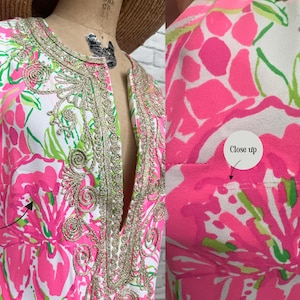 May include: A sleeveless dress featuring a white base with a vibrant pink and green floral pattern. The neckline and front placket are adorned with intricate gold metallic embroidery. The garment includes fine double thread lines as shown.