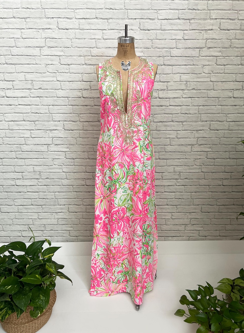 May include: A sleeveless floor-length maxi dress featuring a vibrant pink and lime green floral pattern on a white background. The garment has a deep V-neckline adorned with intricate gold-toned metallic embroidery and beadwork detailing.