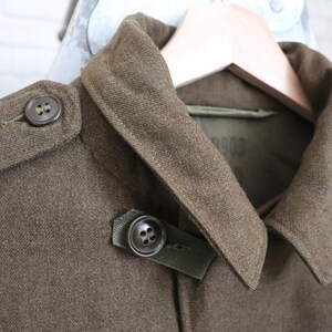 Pu&ograve; includere: Primo piano di una giacca militare verde oliva con colletto a revers e dettagli di bottoni. La giacca &egrave; realizzata in un tessuto strutturato ed &egrave; appesa a una gruccia di legno. I bottoni sono di colore marrone scuro.