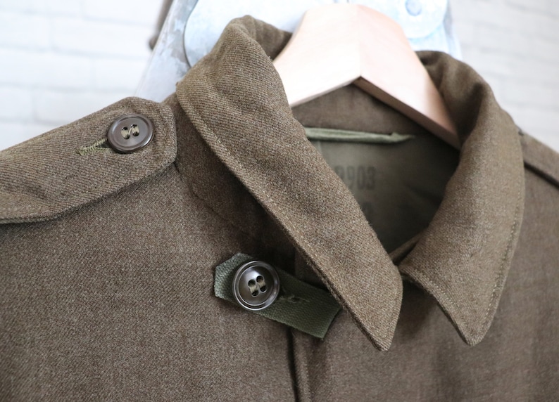 Pu&ograve; includere: Primo piano di una giacca militare verde oliva con colletto a revers e dettagli di bottoni. La giacca &egrave; realizzata in un tessuto strutturato ed &egrave; appesa a una gruccia di legno. I bottoni sono di colore marrone scuro.