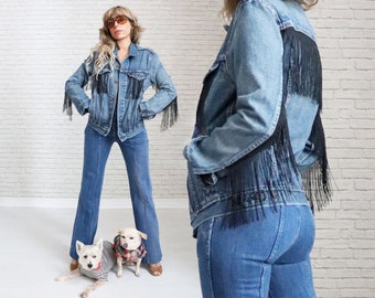 Pode incluir: Uma mulher vestindo uma jaqueta de jeans azul com detalhes de franjas pretas e calças de boca de sino de jeans azul. Ela está em pé com dois cães pequenos brancos vestindo camisas listradas e xadrez.
