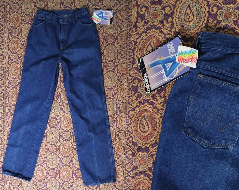 May include: Two pairs of blue denim jeans. One pair is folded and laid flat on a patterned fabric. The other pair is folded and laid flat on a patterned fabric. The jeans have a classic straight leg style. The jeans have a label that says "Wrangler" on it.