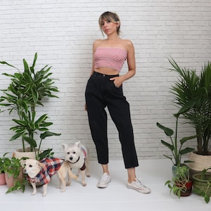 May include: A woman wearing a red and white striped tube top, black high-waisted jeans, and white sneakers. Two small dogs in plaid shirts and a brown vest stand nearby. The background is a white brick wall with potted plants.