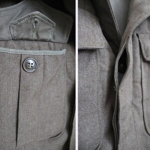 Pu&ograve; includere: Primo piano di una giacca militare verde oliva. La giacca presenta una chiusura con bottoni, due tasche sul petto e una piccola tasca sulla manica superiore sinistra. Il tessuto ha un aspetto strutturato e i bottoni sono grigio scuro.