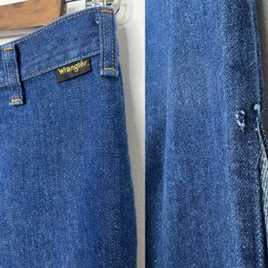 Pode incluir: Um par de cal&ccedil;as de ganga azuis com uma costura dividida na perna direita. As cal&ccedil;as de ganga t&ecirc;m uma etiqueta Wrangler no bolso de tr&aacute;s.