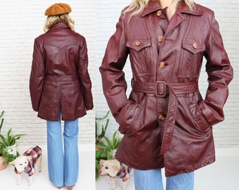 Può includere: Una giacca di pelle bordeaux con cintura e due grandi tasche. La giacca ha una chiusura con bottoni e un colletto.