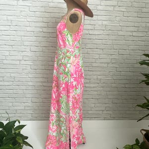 May include: A sleeveless floor-length maxi dress featuring a vibrant floral pattern in shades of hot pink, white, and lime green. The dress has a high neckline and a side slit at the hem. A wide-brimmed straw hat is placed on top of the dress.