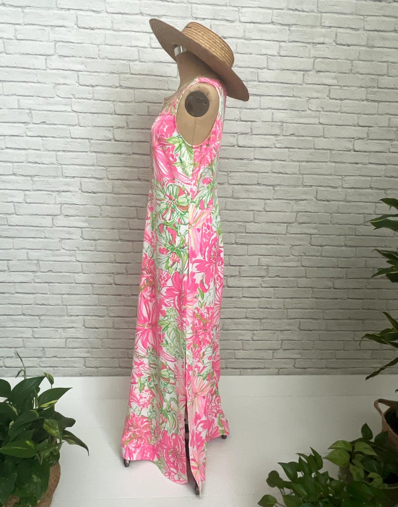May include: A sleeveless floor-length maxi dress featuring a vibrant floral pattern in shades of hot pink, white, and lime green. The dress has a high neckline and a side slit at the hem. A wide-brimmed straw hat is placed on top of the dress.