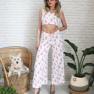 May include: A white floral print two-piece outfit with a cropped top and wide-leg pants. The model is wearing brown heels and sunglasses.