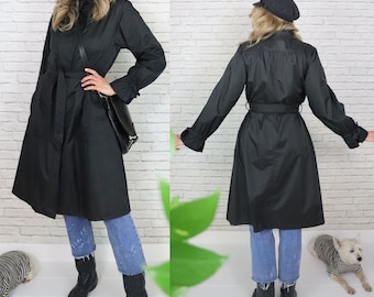 Puede incluir: Una gabardina negra con cinturón y mangas largas. La gabardina tiene un cuello alto y una falda ligeramente acampanada. La modelo lleva pantalones vaqueros azules y botas de vaquero.
