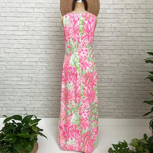 May include: A sleeveless floor-length maxi dress featuring a vibrant floral pattern in shades of hot pink, light pink, and lime green on a white background, displayed on a mannequin wearing a wide-brimmed straw hat.