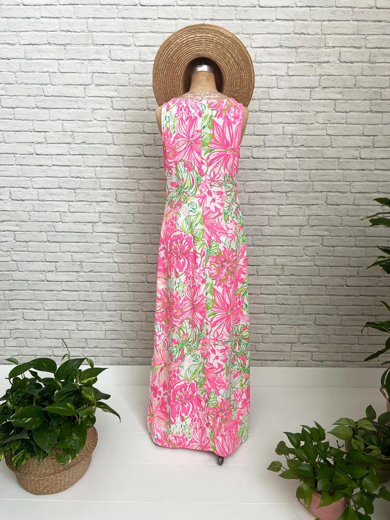 May include: A sleeveless floor-length maxi dress featuring a vibrant floral pattern in shades of hot pink, light pink, and lime green on a white background, displayed on a mannequin wearing a wide-brimmed straw hat.