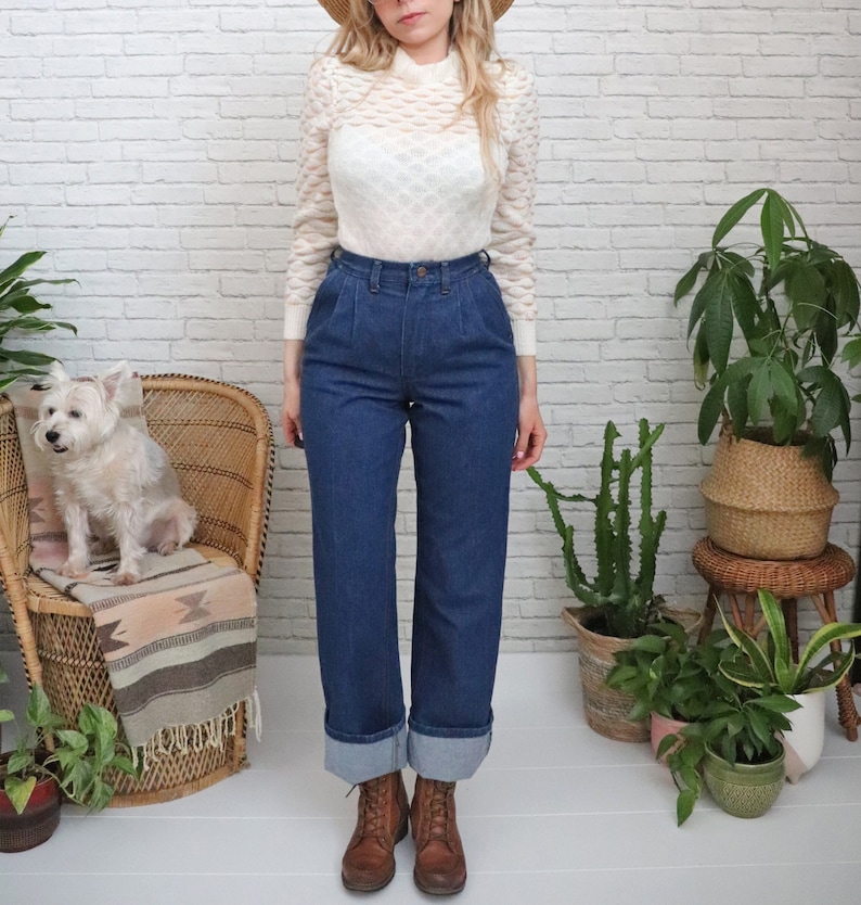 K&ouml;nnte beinhalten: Eine Frau tr&auml;gt einen wei&szlig;en Strickpullover und eine blaue, hochgeschlossene Jeans mit umgeschlagenen S&auml;umen. Sie steht vor einer wei&szlig;en Backsteinwand mit Pflanzen und einem Korbstuhl.