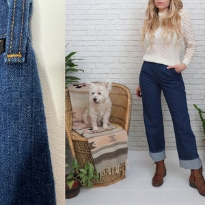 K&ouml;nnte beinhalten: Eine blaue Jeans aus Denim mit umgeschlagenem Saum und einem Wrangler-Etikett. Die Jeans wird von einer Person mit langen blonden Haaren und einem wei&szlig;en Pullover getragen. Die Person steht vor einer wei&szlig;en Backsteinwand.