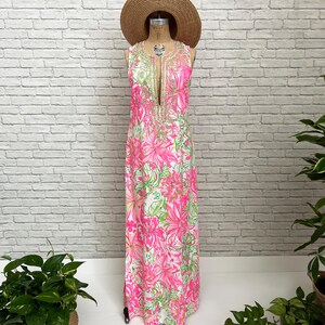 May include: A sleeveless floor-length maxi dress featuring a vibrant floral pattern in shades of hot pink, lime green, and white. The dress has a deep V-neckline adorned with intricate gold-toned embroidery and is displayed on a mannequin with a straw hat.