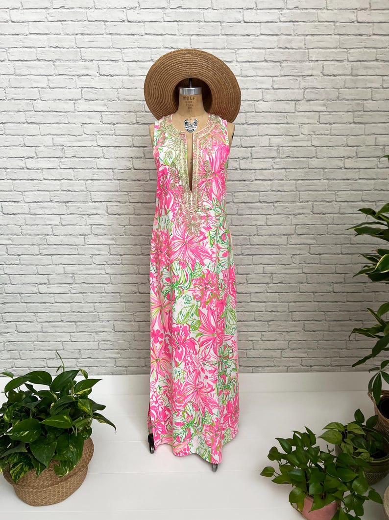 May include: A sleeveless floor-length maxi dress featuring a vibrant floral pattern in shades of hot pink, lime green, and white. The dress has a deep V-neckline adorned with intricate gold-toned embroidery and is displayed on a mannequin with a straw hat.
