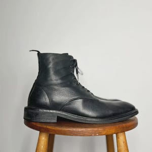 Può includere: Uno stivale in pelle nera con un design stringato. Lo stivale ha una punta arrotondata e un tacco basso. Lo stivale è appoggiato su uno sgabello di legno. Lo stivale è uno stile classico.