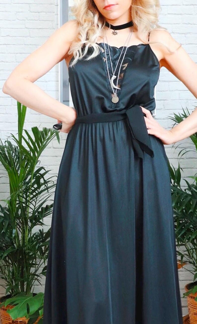 Puede incluir: Un vestido maxi de sat&eacute;n verde oscuro con una faja de sat&eacute;n negro anudada a la cintura. El vestido tiene un escote con ribete de encaje y la modelo lleva varios collares de plata.
