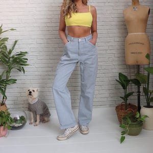 May include: A person wearing a yellow crop top and blue and white striped trousers. The trousers have a high waist and a relaxed fit. White trainers complete the outfit. A small dog in a striped top sits nearby.