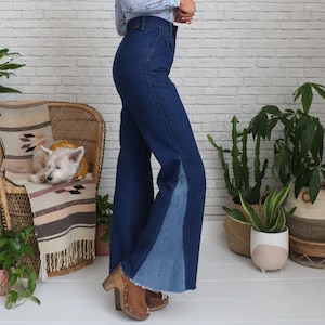 Pode incluir: Uma mulher vestindo um par de cal&ccedil;as jeans de sino azul de denim com um painel azul contrastante na parte inferior de cada perna. As cal&ccedil;as jeans t&ecirc;m cintura alta e um caimento relaxado.